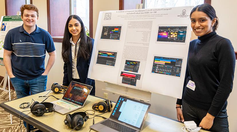 Tanvi, Yasmine, & Joseph stand next to their poster and a table with laptops and headphones.